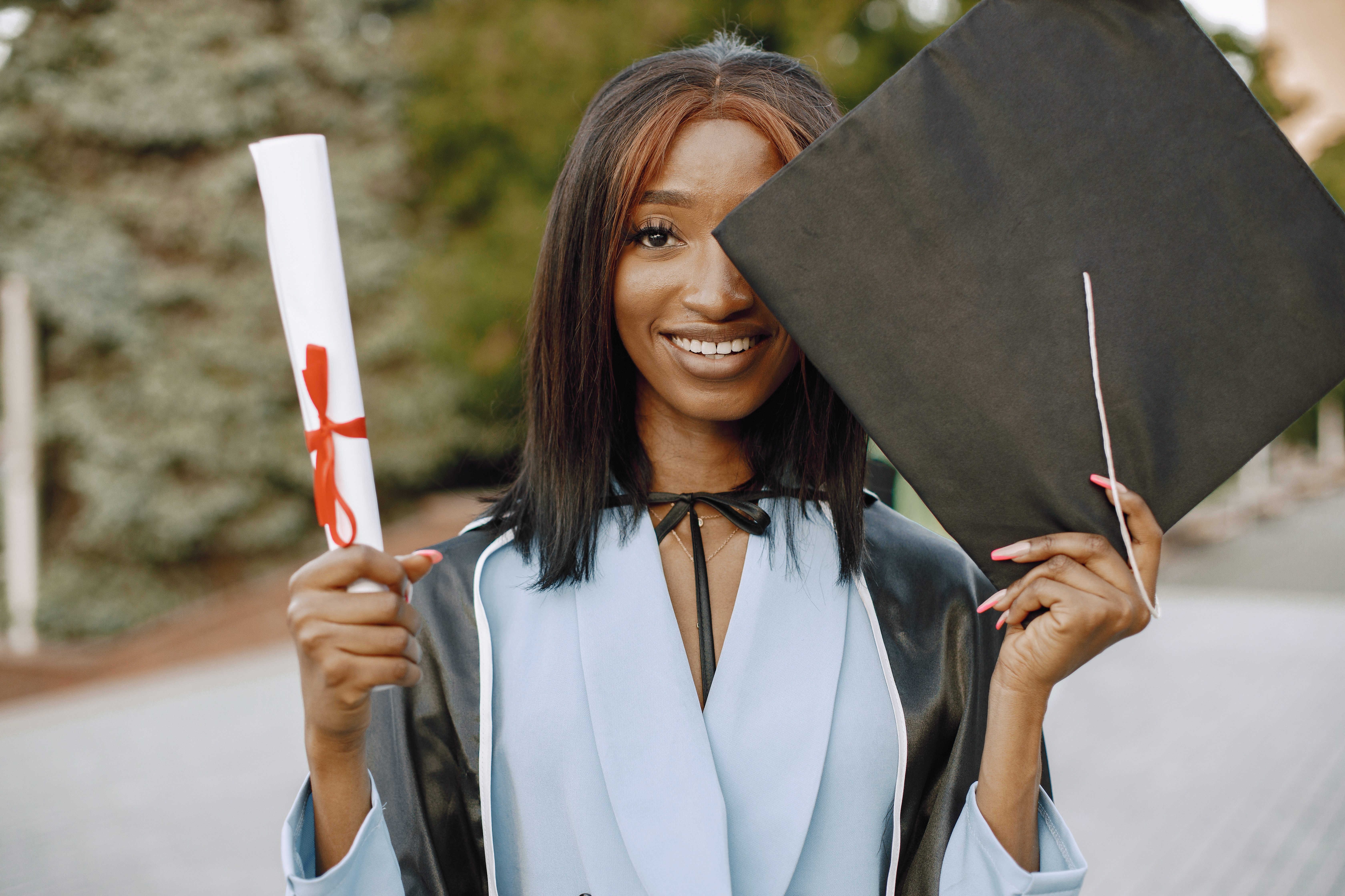 Young afro american female student dressed in black graduation gown. Campus as a background. Girl cheerfully smiling with arms up, holding diploma and academic cap.