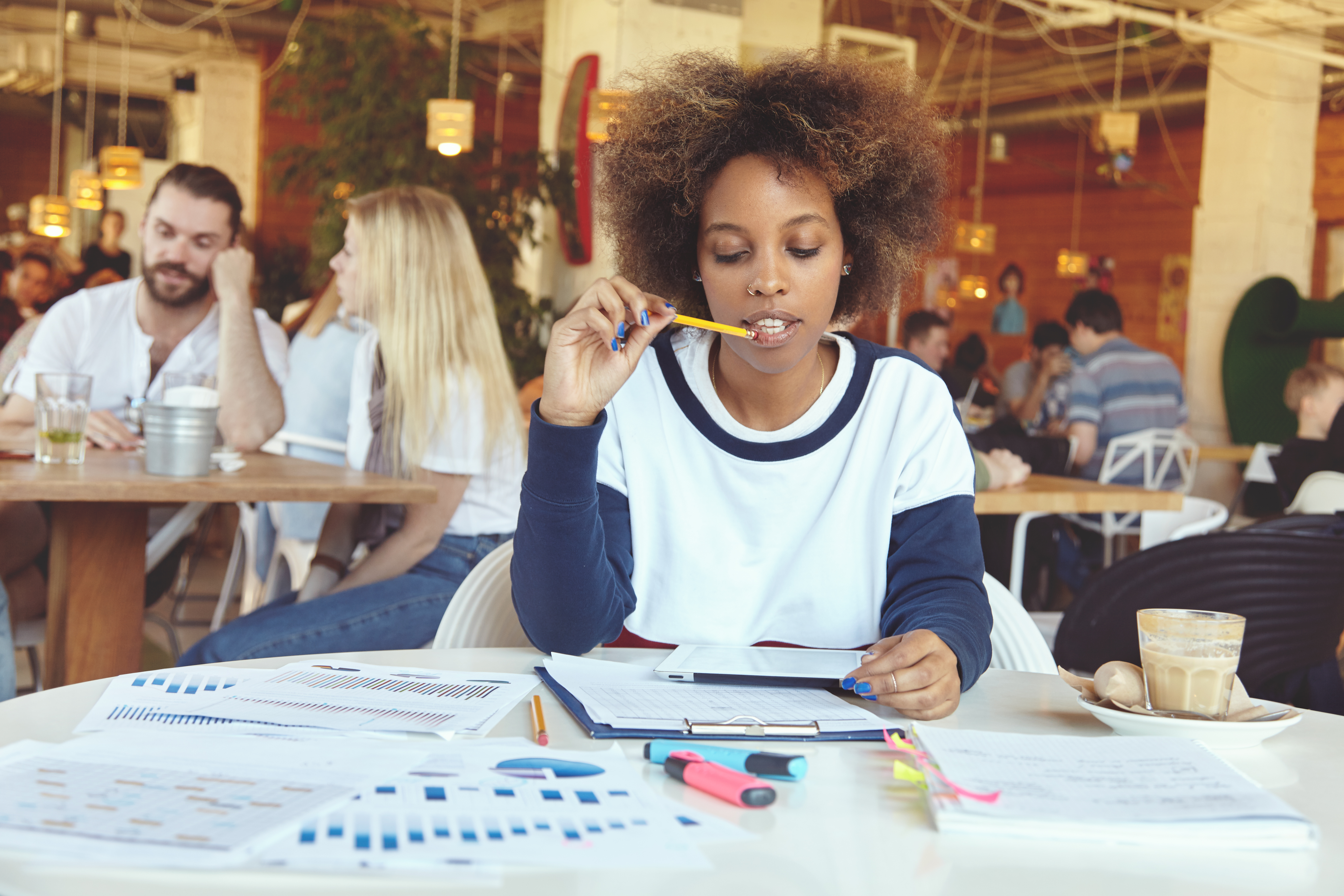 Attractive African student girl in casual clothes sitting at university canteen with touch pad pc, surfing Internet while preparing for exams, touching her lips with pencil, looking thoughtful