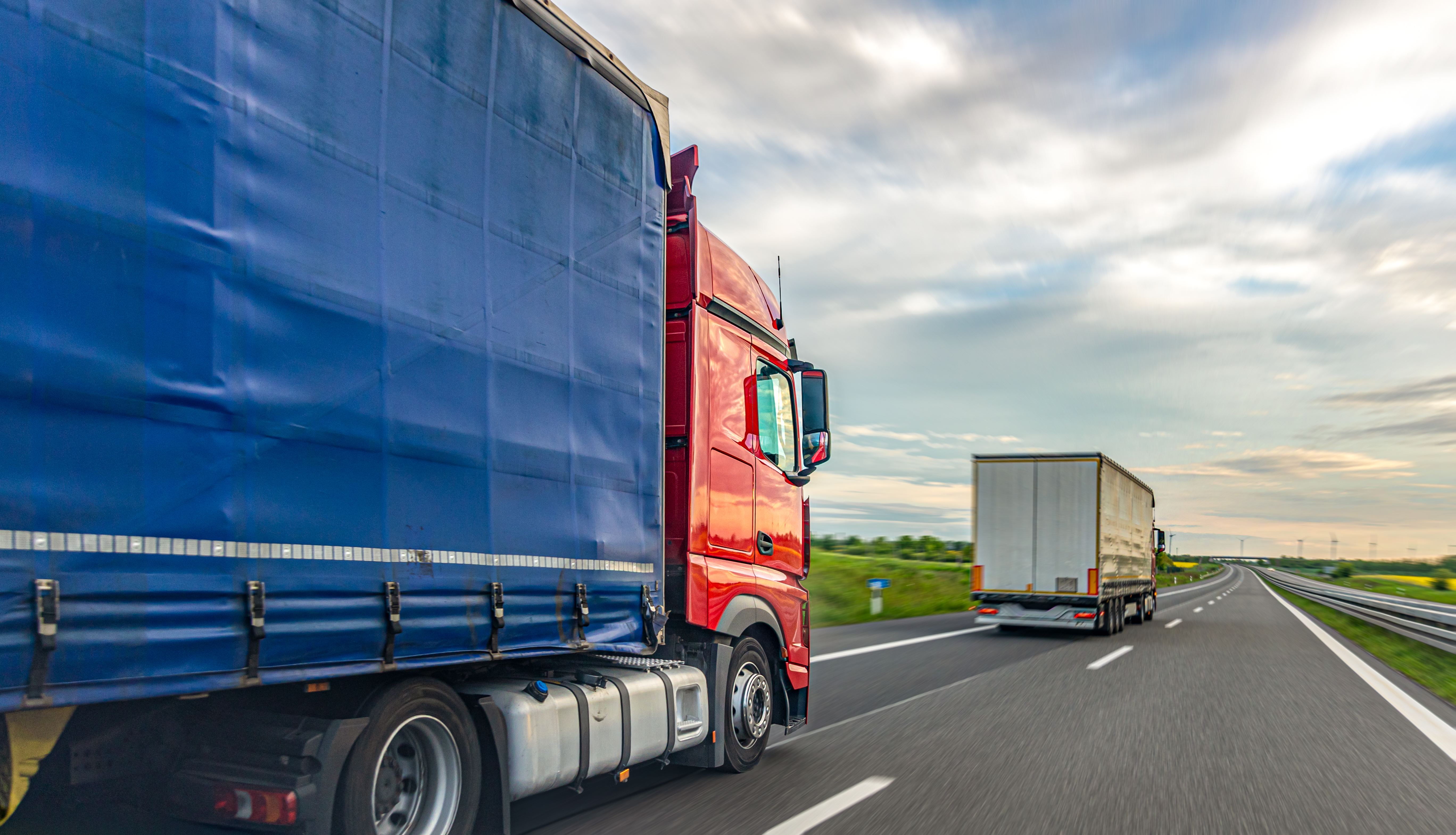 A red and blue freight truck drives along a highway beside another truck under a cloudy sky. Useful for logistics websites, supply chain articles, freight transport content, and industry presentations.
