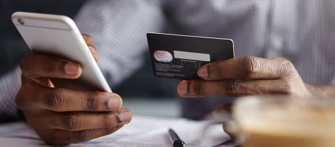 Cropped shot of African-American businessman paying with credit card online making orders via Internet. Successful black male holding plastic card making transaction using mobile banking application