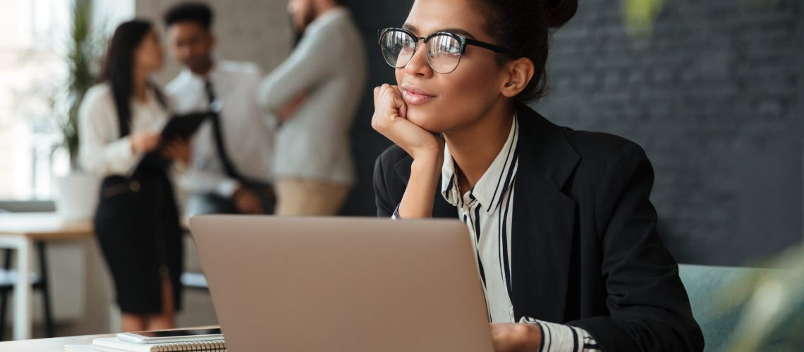 Photo of dreaming young african business woman sitting indoors using laptop computer. Looking aside.