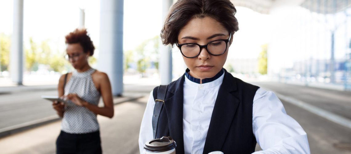 Young successful businesswoman in glasses looking at watch, holding coffee, standing near business centre.